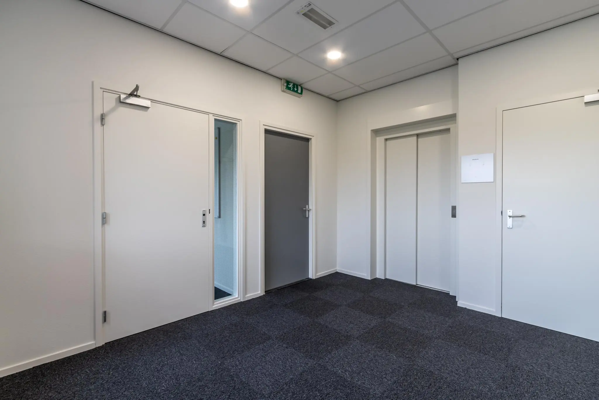 Moderne hal met grijze tapijttegels, meerdere deuren en een lift in een kantoorpand aan de Lage Biezenweg.